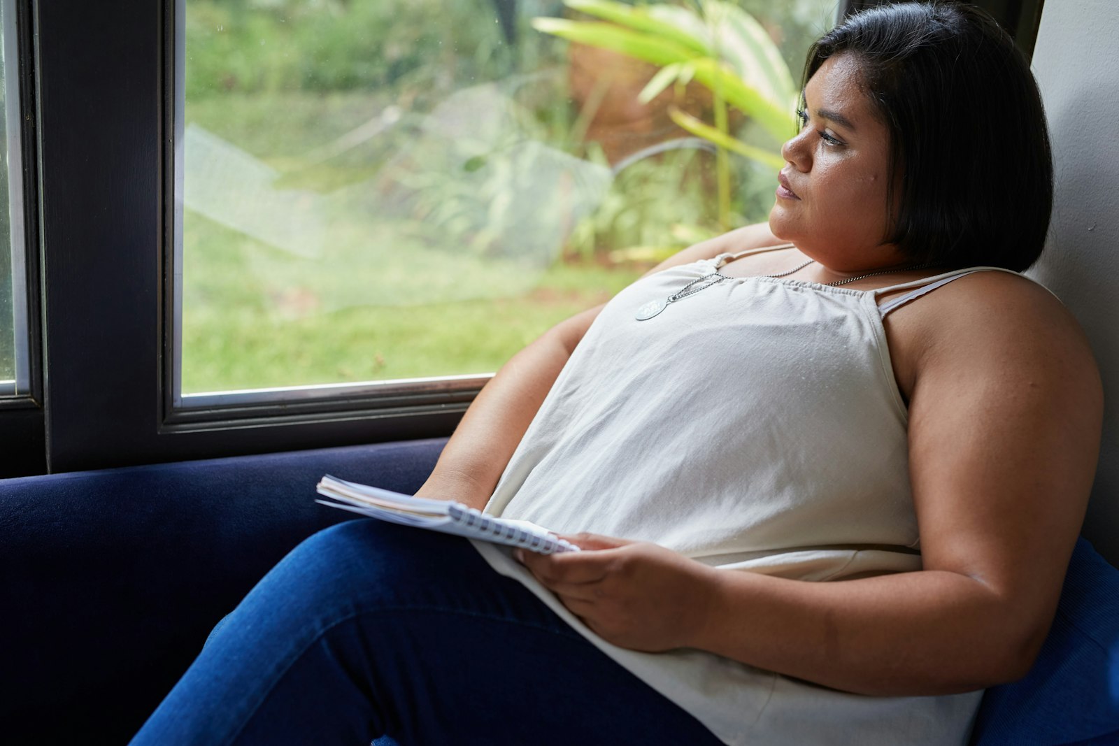 A woman sitting quietly by a window with a journal in her lap, gazing out into soft natural light as she pauses to notice what she is feeling, representing the reflective work of Emotionally Focused Individual Therapy